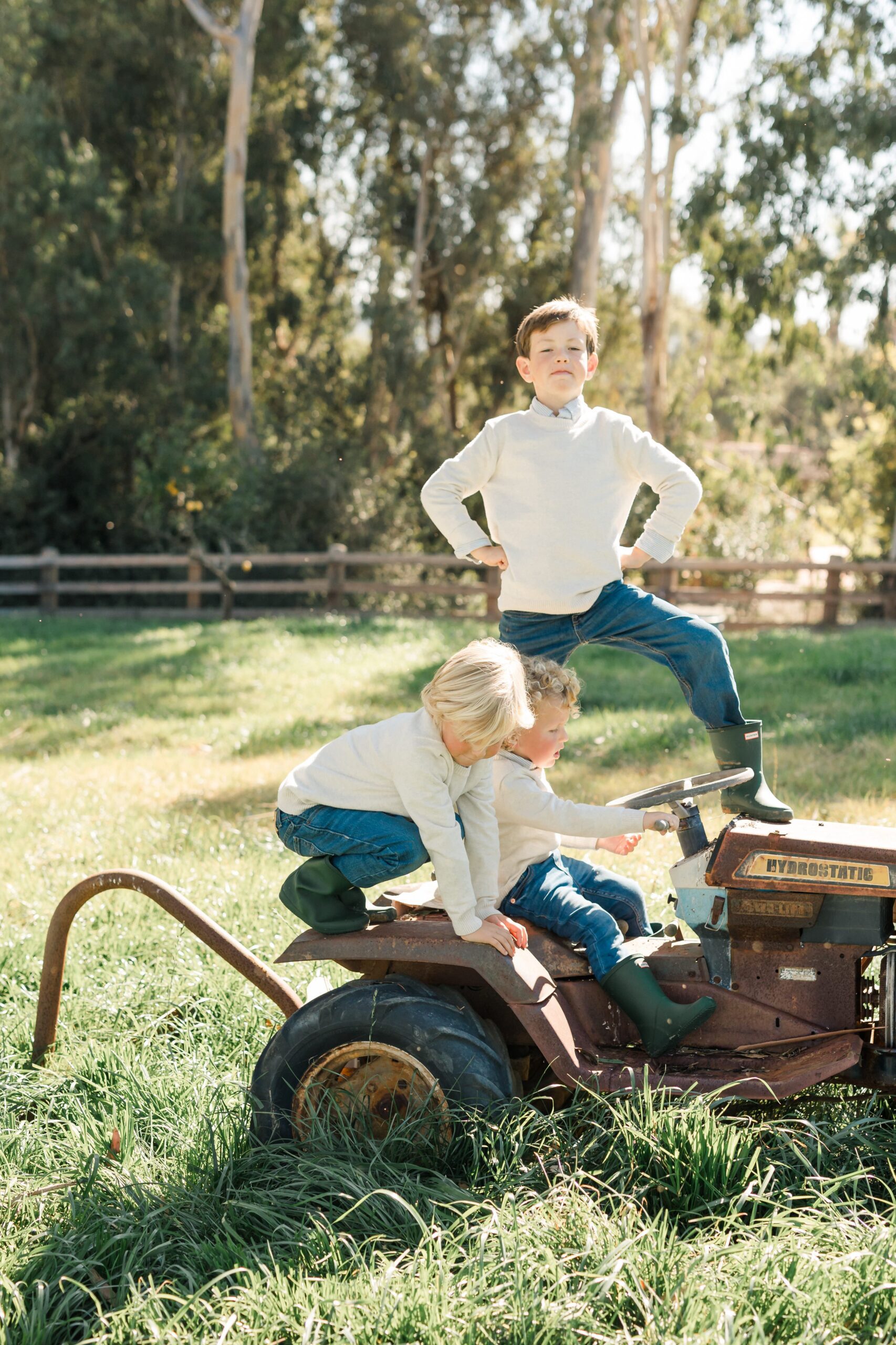 Scott Fell HIGH REZ-202 Three children play on a vintage tractor in a grassy field. One stands on the seat, while the other two climb on the front. Trees and a wooden fence are in the background.